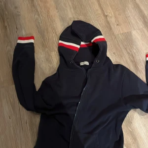 Mörkblå huvtröja från Moncler - Säljer en snygg mörkblå huvtröja från Moncler med dragkedja och ränder i rött och vitt på ärmarna och huvan. Perfekt för en stilren look. Tröjan har en broderad Moncler-logga på ärmen.