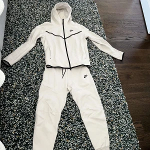 Vit träningsdräkt från Nike - Snygg vit träningsdräkt från Nike med dragkedja och huva. Jackan har svarta detaljer och byxorna har elastisk midja. Perfekt för träning eller en avslappnad dag.