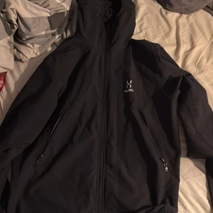 Svart jacka från Haglöfs - Säljer en svart jacka från Haglöfs med dragkedja och huva. Jackan har två sidofickor med dragkedjor och är perfekt för utomhusaktiviteter. Den är stilren och funktionell, idealisk för både stad och natur. Lite skadad nere vid midjan men inget märkvärdigt därav priset 