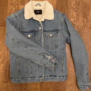 Jeansjacka med teddyfoder från Bik Bok - Snygg jeansjacka från Bik Bok med mjukt teddyfoder och klassisk krage. Jackan har knappar framtill och två bröstfickor med lock. Perfekt för kyligare dagar och ger en avslappnad stil.