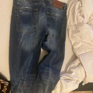 Blå jeans från Dsquared2 - Snygga blå jeans från Dsquared2 med slitna detaljer och färgstänk. De har en klassisk femficksdesign och en knappgylf. Perfekta för en avslappnad och trendig look. Priset kan diskuteras 