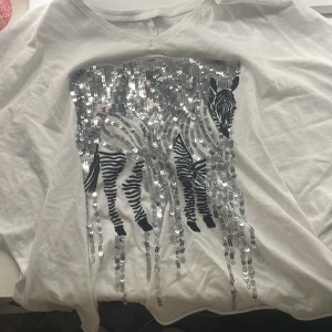 Vit kortärmad topp med zebramönster och paljetter - Snygg vit kortärmad topp med ett coolt zebramönster i silverpaljetter på framsidan. Perfekt för att lägga till lite glitter i vardagen. Toppen har en bekväm passform och en rund halsringning. Sitter overzist.