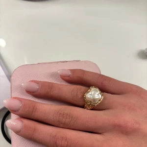 Guldfärgad ring med pärldetalj - Elegant guldfärgad ring med en stor pärldetalj i mitten. Ringen har en vacker, detaljerad design som ger ett lyxigt intryck. Perfekt för att ge en touch av glamour till din stil.