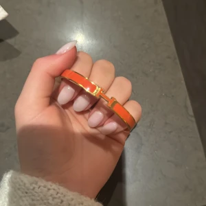 Orange armband med gulddetaljer - Snyggt orange armband med gulddetaljer och ett ikoniskt H-spänne. Perfekt för att ge en färgklick till din outfit. Armbandet har en elegant och stilren design. Lite svårt att knäppa det det är trasigt sen jag köpte det så de åker av lätt men säkert enkelt att laga