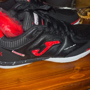 Svarta sneakers från Joma - Snygga svarta sneakers från Joma med röda detaljer och vit sula. Skorna har snörning och en sportig design, perfekt för en aktiv livsstil.