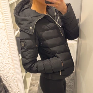 Svart dunjacka från Hollies - Nypris 3000-4500kr. Snygg svart dunjacka från Hollies med dragkedja och flera fickor. Jackan har en huva och en liten logotyp på ärmen. Den har många fina detaljer och är i nyskick. 
