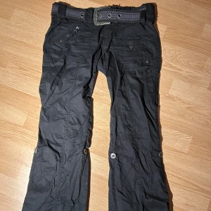 Svarta cargopants med bälte - Snygga svarta cargopants med flera fickor och ett matchande bälte. Byxorna har knappar och dragkedja samt en cool design med detaljerade sömmar. 