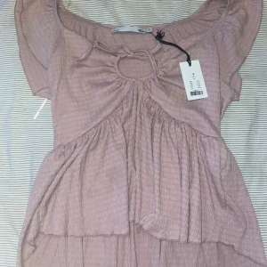 Ljusrosa topp med volanger - Säljer en superfin ljusrosa topp med volanger och en söt knytdetalj framtill. Toppen har en offshoulder-design och är perfekt för sommardagar. Materialet är lätt och luftigt, vilket gör den bekväm att bära. Den är i nyskick och har fortfarande prislappen kvar.