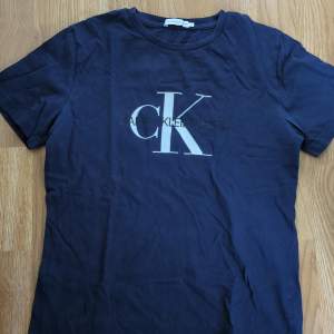 Säljer en snygg mörkblå t-shirt från Calvin Klein. Den har en klassisk passform med korta ärmar och ett stort CK-logotryck på framsidan. Perfekt för en casual look eller att matcha med jeans. T-shirten är i bra skick och redo för en ny ägare!