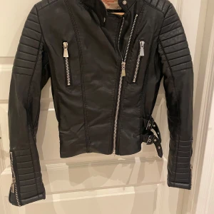 Svart skinnjacka med dragkedjor - Säljer en snygg svart skinnjacka med coola dragkedjor och bältesdetaljer. Jackan har en tight passform och långa ärmar med quiltade detaljer på axlarna. Perfekt för höst och vår!