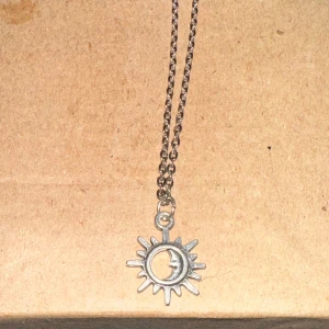 Silverfärgat halsband med sol- och månhänge - Säljer ett snyggt silverfärgat halsband med ett unikt hänge som föreställer en sol med en måne inuti. Perfekt för att ge en mystisk touch till din outfit. Kedjan är tunn och elegant, vilket gör det lätt att bära till både vardags och fest.