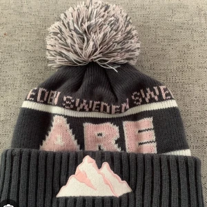 Åre mössa - Snygg grå mössa med stor tofs och ett coolt bergsmotiv framtill. Mössan har text i rosa och vitt samt ribbad kant. Perfekt för kyliga dagar!