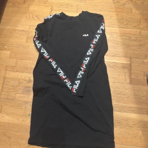 Svart långärmad klänning från FILA - Säljer en svart långärmad klänning från FILA med logotyp längs ärmarna i vitt och rött. Perfekt för en sportig look.