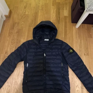 Mörkblå dunjacka från Stone Island - Snygg mörkblå dunjacka från Stone Island med luva och dragkedja. Jackan har det ikoniska Stone Island-märket på ärmen och praktiska fickor med dragkedjor. Perfekt för kyligare dagar. Storlek XL. pris kan diskuteras och tar byten. Nypris 6400.