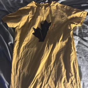 Gul t-shirt från Dsquared2 - Säljer en gul t-shirt från Dsquared2 med ett svart grafiskt tryck på framsidan. T-shirten är kortärmad och tillverkad i bomull, perfekt för en avslappnad stil till sommaren för att fånga blickarna med den synliga färgen perfekt somma tröja.