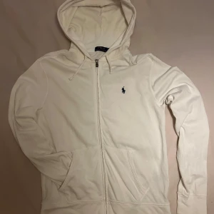 Vit hoodie från Ralph Lauren - Säljer en stilren vit hoodie från Ralph Lauren med dragkedja och klassisk logga på bröstet. Perfekt för en avslappnad look. Storlek är M och nypris är (1699kr)
