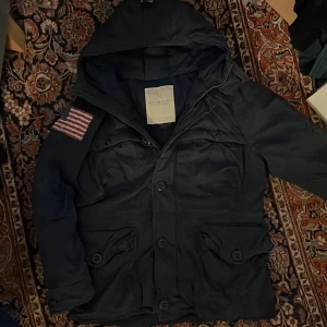 Mörkblå vinterjacka från Denim & Supply - Snygg mörkblå jacka från Denim & Supply av Ralph Lauren. Jackan har en huva och en cool amerikansk flagga på ärmen. Den stängs med både dragkedja och knappar, vilket ger en stilren look. Perfekt för vintern eftersom den är väldigt fodrar