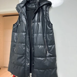 Svart quiltad väst från Zara - Snygg svart quiltad väst från Zara med dragkedja och knappar. Perfekt för lager-på-lager-stil med huva för extra värme. Västen har en modern look och är idealisk för kyligare dagar. Passar perfekt s/m/l storlek