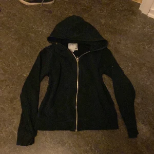 Svart huvtröja med dragkedja - Säljer en svart huvtröja med dragkedja framtill. Tröjan har långa ärmar och en praktisk huva, perfekt för kyliga dagar. Enkel och stilren design som passar till det mesta.