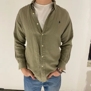 Olivgrön skjorta från Ralph Lauren - Olivgrön skjorta från Ralph Lauren med klassisk krage och knappar framtill. Skjortan har långa ärmar och en liten broderad logotyp på bröstet. Den är gjord av 51% silke och 49% linne. 