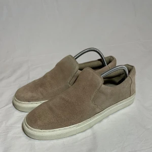 Beige mocka slip-on skor från Jack & Jones - Snygga beige slip-on skor i mocka från Jack & Jones. Skorna har en vit sula som ger en fräsch kontrast till den mjuka mockan. Storleken är 42 och de är använda många gånger. 