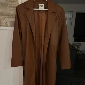Brun trenchcoat från Zara - Snygg brun trenchcoat från Zara i syntetmaterial. Jackan har en klassisk design med knytbälte i midjan och långa ärmar. Perfekt för höst och vår.