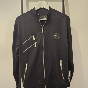 Philipp Plein kofta  - Snygg svart bomberjacka från Philipp Plein med dragkedjor och ett coolt dödskallemotiv på ryggen. Jackan har flera dragkedjedetaljer framtill och en rund halsringning. Helt ny