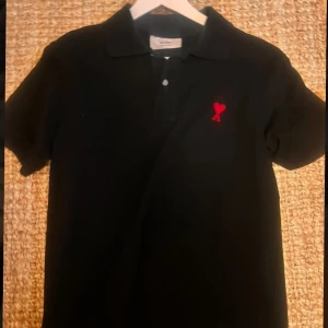 Ami Paris pike - Snygg svart pikétröja från Ami Paris med en röd broderad detalj på bröstet. Tröjan har en klassisk krage och knappar framtill. Perfekt för en stilren look. Tar byten 