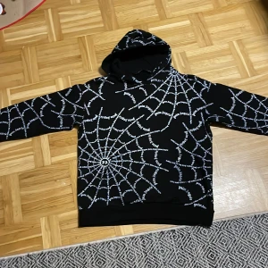 Svart hoodie med spindelnätsmönster från Palm Angels - Säljer en cool svart hoodie från Palm Angels med ett unikt spindelnätsmönster i vitt. Hoodien har en klassisk passform med långa ärmar och en bekväm huva. Perfekt för dig som vill sticka ut!