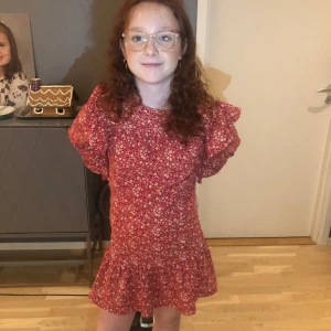 Röd blommig klänning med volangdetaljer - Charmig röd klänning med blommigt mönster och volangdetaljer. Klänningen har långa ärmar och en kort längd, perfekt för en somrig look. Det står att det är storlek 140 men den  passade min kompis perfekt och hon har 146 till 152