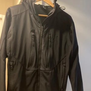 Svart jacka från RevolutionRace - Snygg svart jacka från RevolutionRace med dragkedja och huva. Perfekt för utomhusaktiviteter med flera praktiska fickor. Designad i Sverige . 