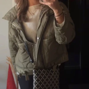 Olivgrön quiltad jacka från Zara - Säljer en olivgrön quiltad jacka från Zara med dragkedja och knappar. Jackan har långa ärmar och praktiska fickor framtill. Perfekt för kyligare dagar.