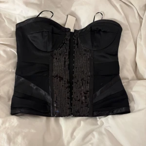 Svart korsett med paljetter - Snygg svart korsett med glittrande paljettdetaljer framtill. Superfin! Har tyvärr tappat några paljetter (se bild) annars så har den prislappen på! Nypris 35€ passform som framhäver figuren. Perfekt för en utekväll eller fest. Passar även till nyår!