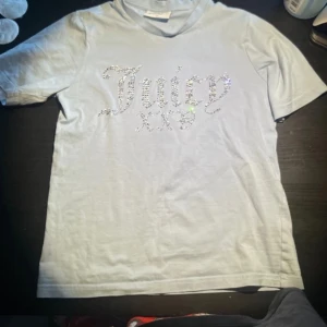 Juicy couture ljus blå t shirt - Snygg ljusblå t-shirt från Juicy Couture med glittrande stenar som bildar texten 'Juicy' på framsidan. Perfekt för en trendig och avslappnad stil.