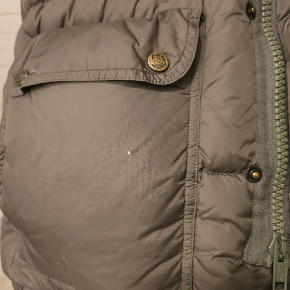Säljer en grön dunjacka, expedition down från Fjällräven med huva och gula detaljer. Jackan har en broderad logotyp på bröstet och praktiska fickor med knappar. Perfekt för kyliga dagar. Jackan är storlek xs och personen på bilden är 166 och har storlek xxs i kläder. Finns ett litet hål på ena fickan, se bilder. . Takit.