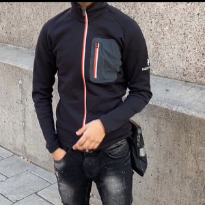 Svart  Peak Performance kofta  - Snygg svart kofta  från Peak Performance med orange dragkedja och en praktisk bröstficka. Perfekt för en sportig look och passar bra till vardagsäventyr.