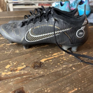 Nike mercurial vapor 14 elite svarta, storlek 39 - Snygga svarta fotbollsskor från Nike med gulddetaljer och snörning. Skorna har en slitstark sula med dobbar för bra grepp på planen. Perfekta för fotbollsspelare som vill ha stil och funktion. 8/10 i betyg. Använda i 2 månader och köpta för 2800 kr. 