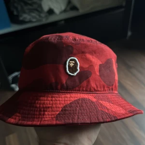 Bape fiskehatt - Snygg röd kamouflagehatt från A Bathing Ape med broderad logotyp framtill. Perfekt för att ge din outfit en cool touch. Hatten har en klassisk bucket-modell och är tillverkad i bomull.