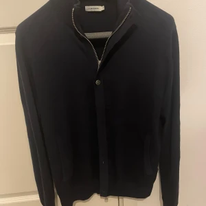Mörkblå kofta från J.Lindeberg - Snygg mörkblå kofta/ cardigan/ fullzip från J.Lindeberg med dragkedja och knappdetalj. Perfekt för kyligare dagar med sin stilrena design och långa ärmar. Passar både till vardags och mer uppklädda tillfällen. Knappt använd 