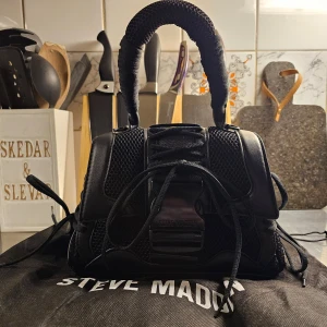 Svart handväska från Steve Madden - Snygg svart handväska från Steve Madden med coola detaljer. Väskan har ett flätat handtag och snördetaljer framtill. Perfekt för att ge din outfit en edgy touch.