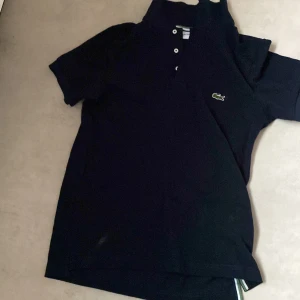 Svart pikétröja från Lacoste - Säljer en klassisk svart pikétröja från Lacoste med korta ärmar och den ikoniska krokodilloggan på bröstet. Tröjan har en knappslå med tre knappar och en ribbad krage med en grön och vit rand.