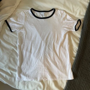 Vit t-shirt med svarta kanter  - Säljer en stilren vit t-shirt från primark med svarta kanter runt halsen och ärmsluten. Perfekt för en avslappnad look. T-shirten är i bomull och har en normal passform. Passar till både vardag och fest!