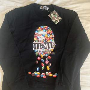 Säljer en svart tröja från A Bathing Ape med ett färgglatt M&M's motiv på framsidan. Tröjan har långa ärmar och är i nyskick. Perfekt för dig som vill ha något unikt och stilrent i garderoben. Taggarna sitter fortfarande kvar!