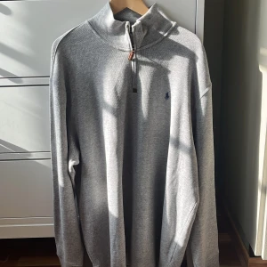 Grå tröja med dragkedja från Polo Ralph Lauren - Säljer en stilren grå tröja från Polo Ralph Lauren i storlek XXL. Tröjan har en halv dragkedja och är långärmad, perfekt för både höst och vinter. Den är gjord i ett mjukt material som känns skönt mot huden. En liten broderad logga på bröstet ger en extra touch av stil. Passar både till vardags och lite mer uppklädda tillfällen.