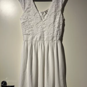 Vit spetsklänning - Säljer en superfin vit klänning med spetsdetaljer. Den har en vacker spetsöverdel och en kjol som faller fint. Perfekt för sommarens fester eller kanske till studenten. Klänningen har en dragkedja i ryggen och är ärmlös för en elegant look.