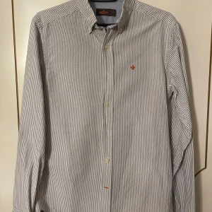 Randig skjorta från Morris - Säljer en stilren och klassisk randig skjorta från Morris i storlek S. Skjortan har en snygg vit och blå randig design med en liten orange broderad detalj på bröstet. Nypris 1700