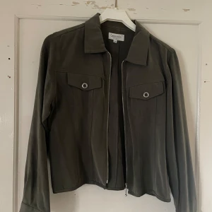 Grön jacka från Cappuccini - Säljer en snygg grön jacka från Cappuccini. Den har en stilren design med dragkedja framtill och två fickor med knappar på bröstet. Jackan är långärmad och passar perfekt för både vår och höst. Materialet känns mjukt och bekvämt.