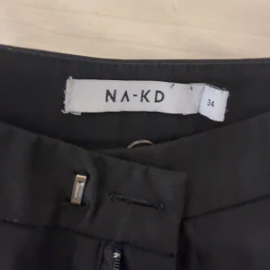 Svarta byxor från NA-KD - Säljer ett par stilrena svarta byxor från NA-KD i storlek 34 alltså nog XS men jag garanterar att dem passar perfekt även för storlek S. De har en klassisk design med dragkedja och knapp framtill. Perfekta för både vardag och fest!