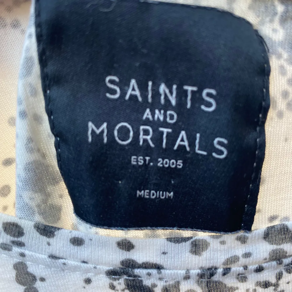 Säljer en cool vit t-shirt från Saints and Mortals med ett svart stänkmönster. Den har korta ärmar och en avslappnad passform, perfekt för en casual look. Materialet känns mjukt och skönt mot huden.. Toppar.