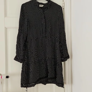Svart prickig klänning från Twist & Tango - Säljer en superfin svart klänning med vita prickar från Twist & Tango. Den har långa ärmar och en lös passform som gör den perfekt för både vardag och fest. Klänningen har en söt volangdetalj längst ner och knappar framtill. Perfekt för höst och vår!